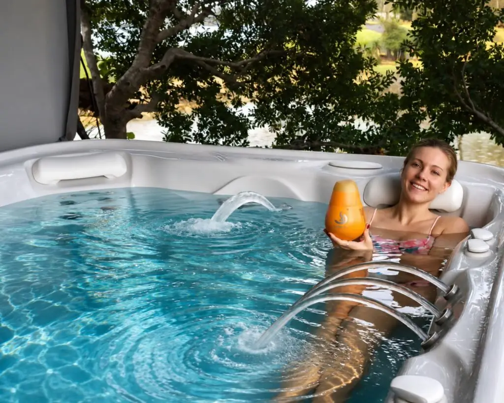 Smiling woman relaxing in an outdoor hot tub, holding an orange bottle amid clear blue water and leafy background.